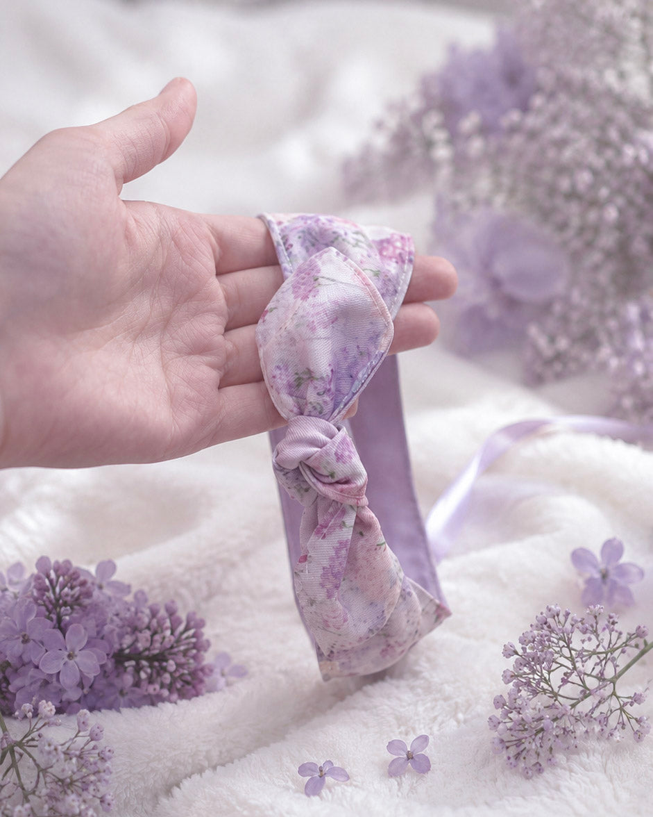 A lavender and white floral satin hair bow held in a person's hand, surrounded by soft white fabric and small purple flowers.