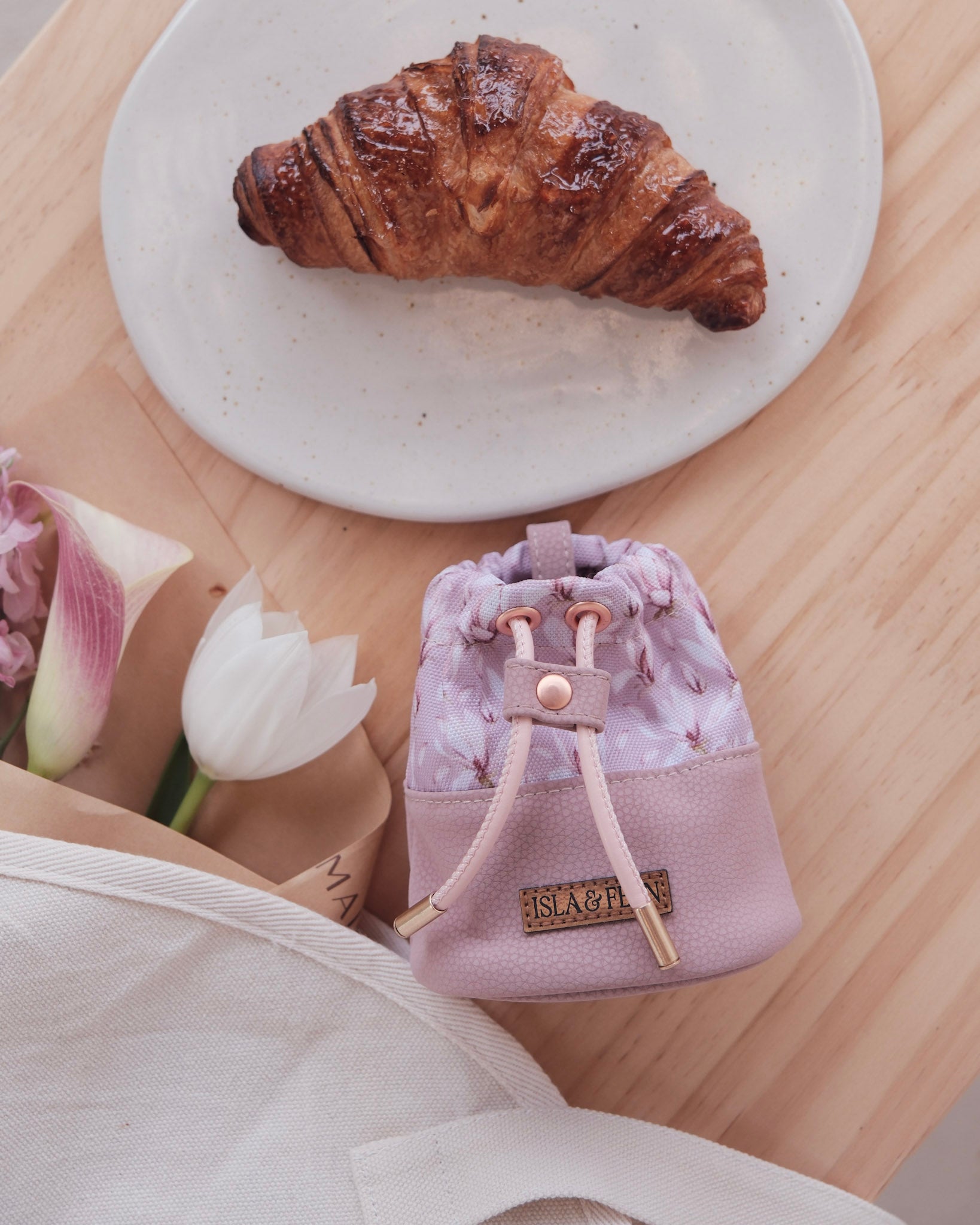 Small purple drawstring bag on a wooden table with a croissant and flowers.