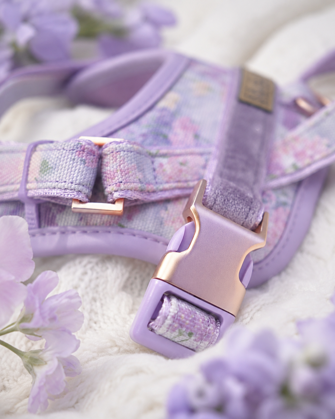 A close-up of a lavender pet harness with a pastel floral pattern, rose gold buckle, and matching lavender collar, styled on a white textured blanket with purple flowers.
