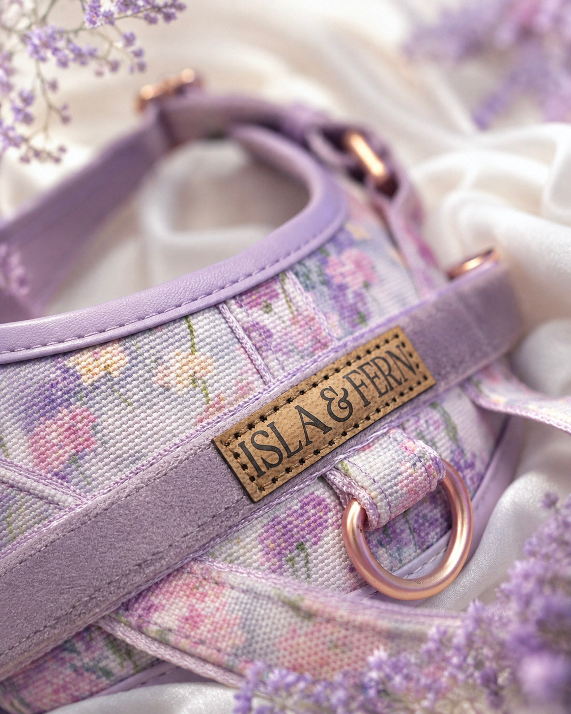 A close-up of a floral-patterned pet harness in lavender and pink tones, featuring a tan leather brand patch that reads 'ISLA & FERN' and a rose gold metal ring, styled with white fabric and lavender flowers.