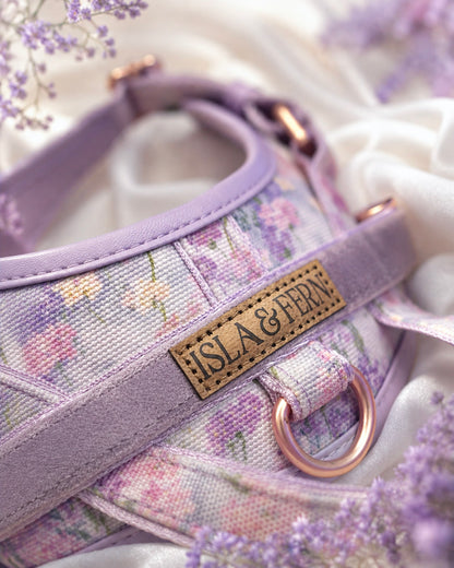 A close-up of a floral-patterned pet harness in lavender and pink tones, featuring a tan leather brand patch that reads 'ISLA & FERN' and a rose gold metal ring, styled with white fabric and lavender flowers.
