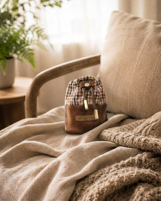A small brown leather and houndstooth drawstring pouch bag with a gold-tone clasp sits on a beige throw blanket draped over a wooden armchair in a softly lit room.