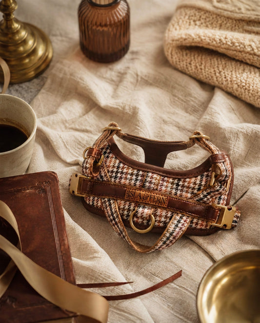 A small houndstooth shoulder bag with brown leather trim and gold-tone hardware, styled on a beige linen surface with a coffee cup, notebook, ribbon, and knit fabric in the background.